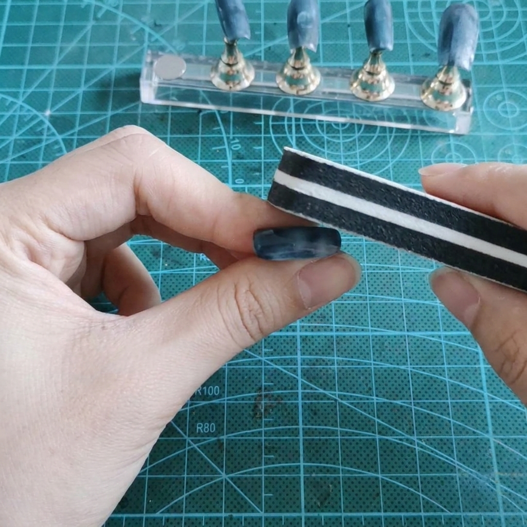 A block sponge buffer sanding down the shiny gel surface of a false nail held between fingers.