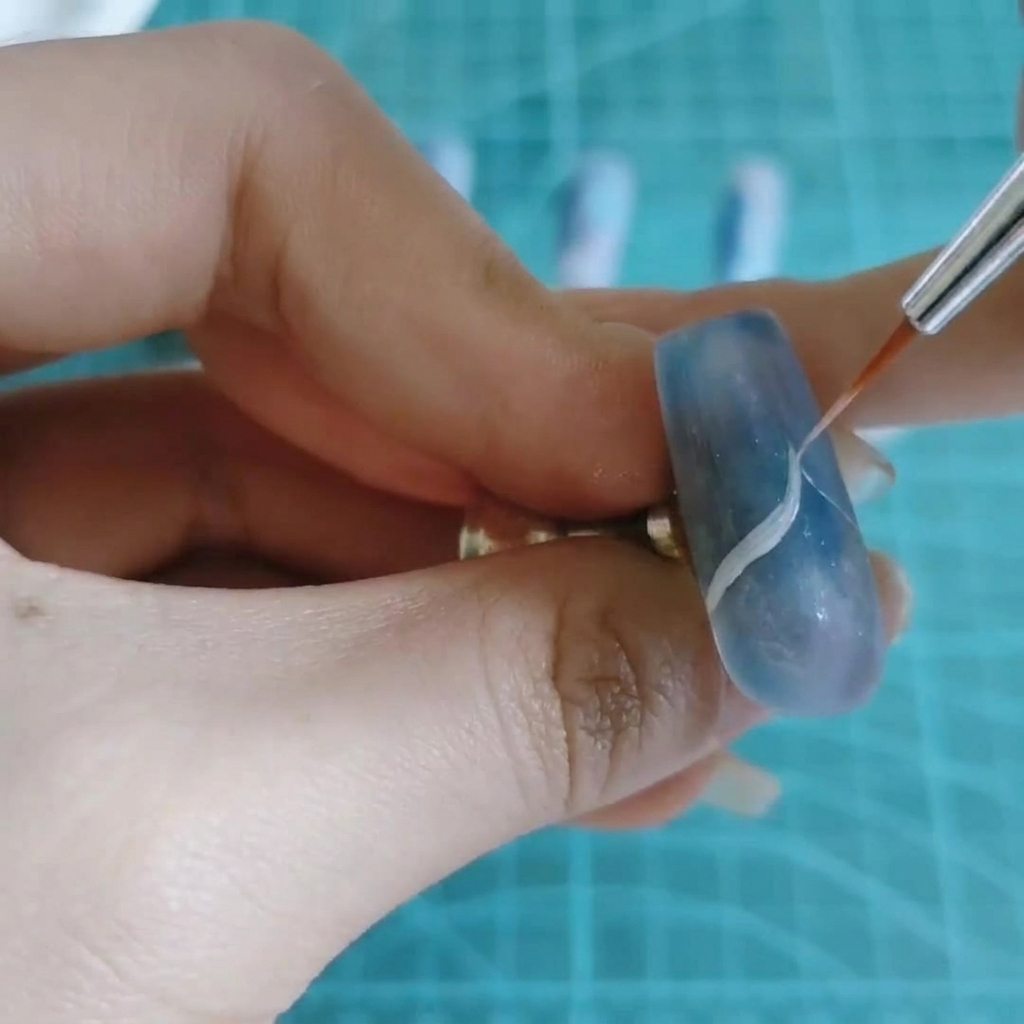 A fine nail art brush drawing a thin white wavy line across a blue and purple marbled nail.