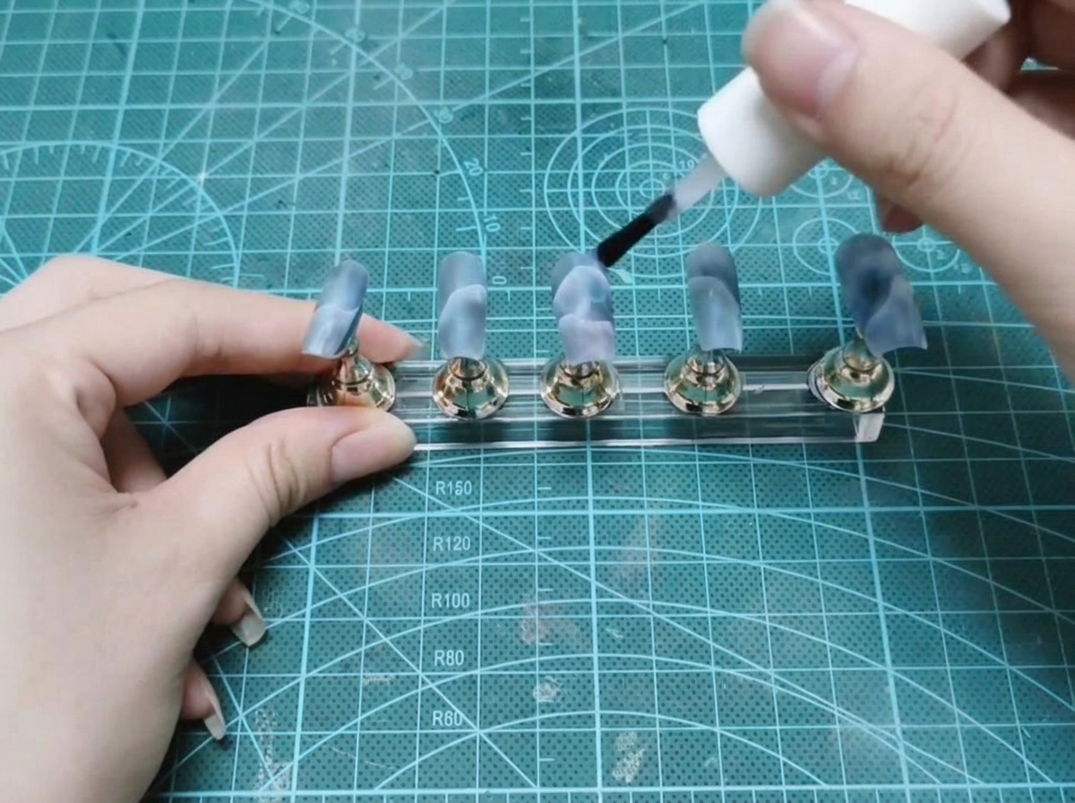 A bottle brush applying clear builder gel to a set of painted false nails on a display stand.