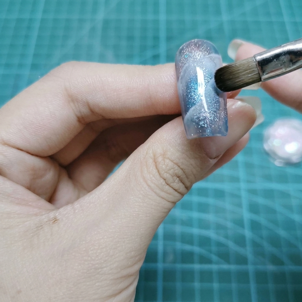 A flat makeup brush tapping holographic glitter particles onto a wet gel nail surface.