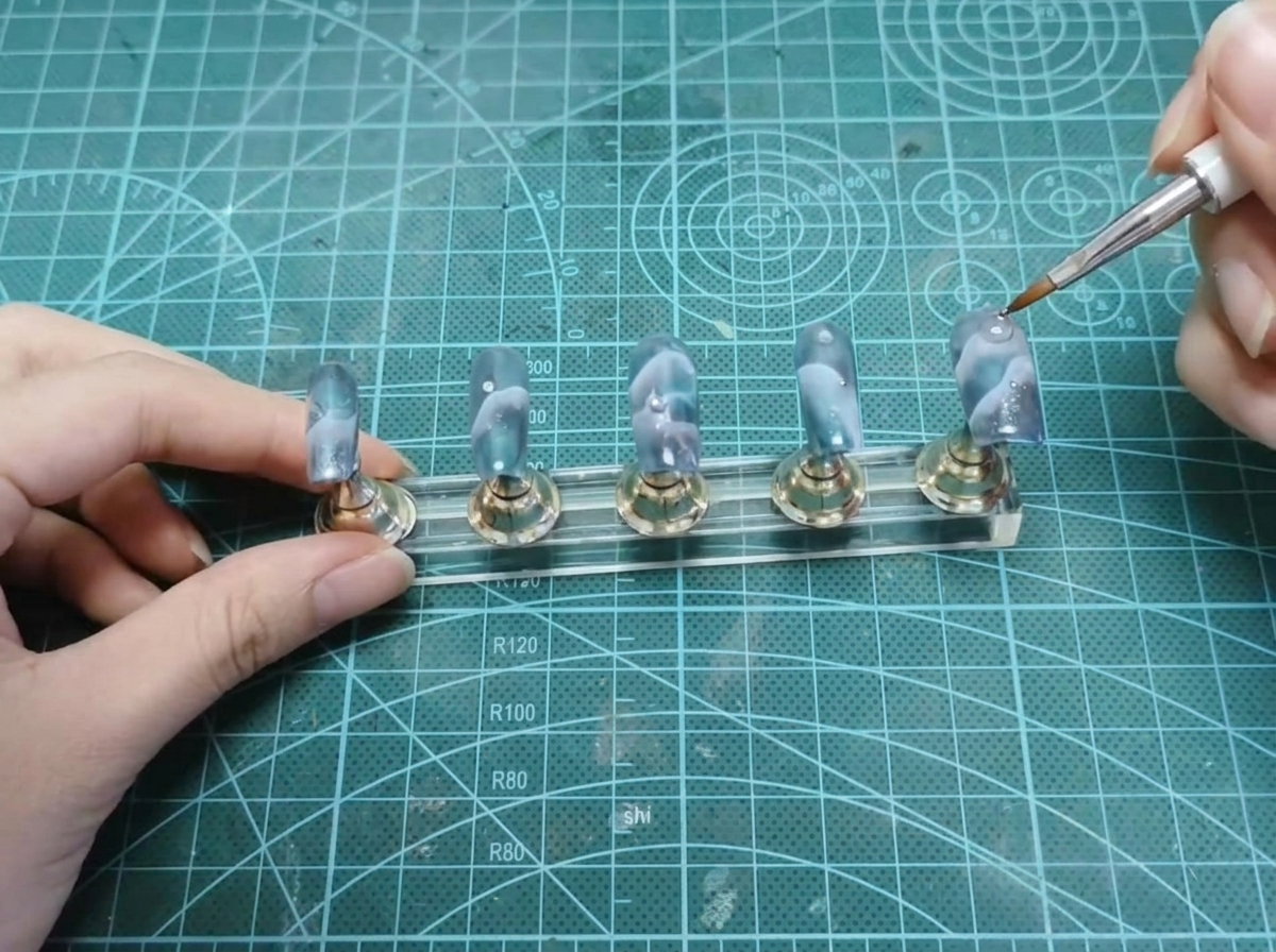 A dotting tool placing a tiny silver bead near the cuticle area of a nail on a stand.