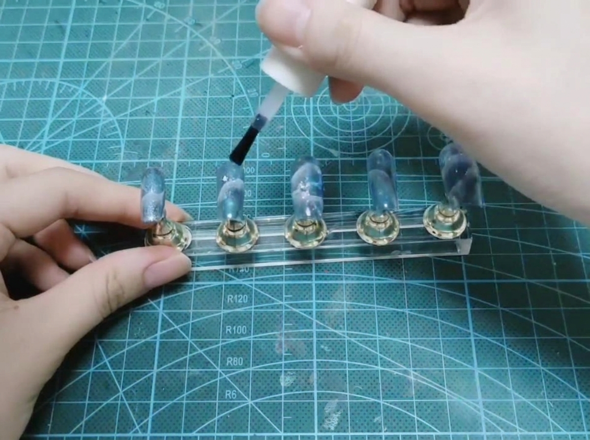 A hand using a brush from a white bottle to apply glossy top coat over a set of completed nails on a display stand.