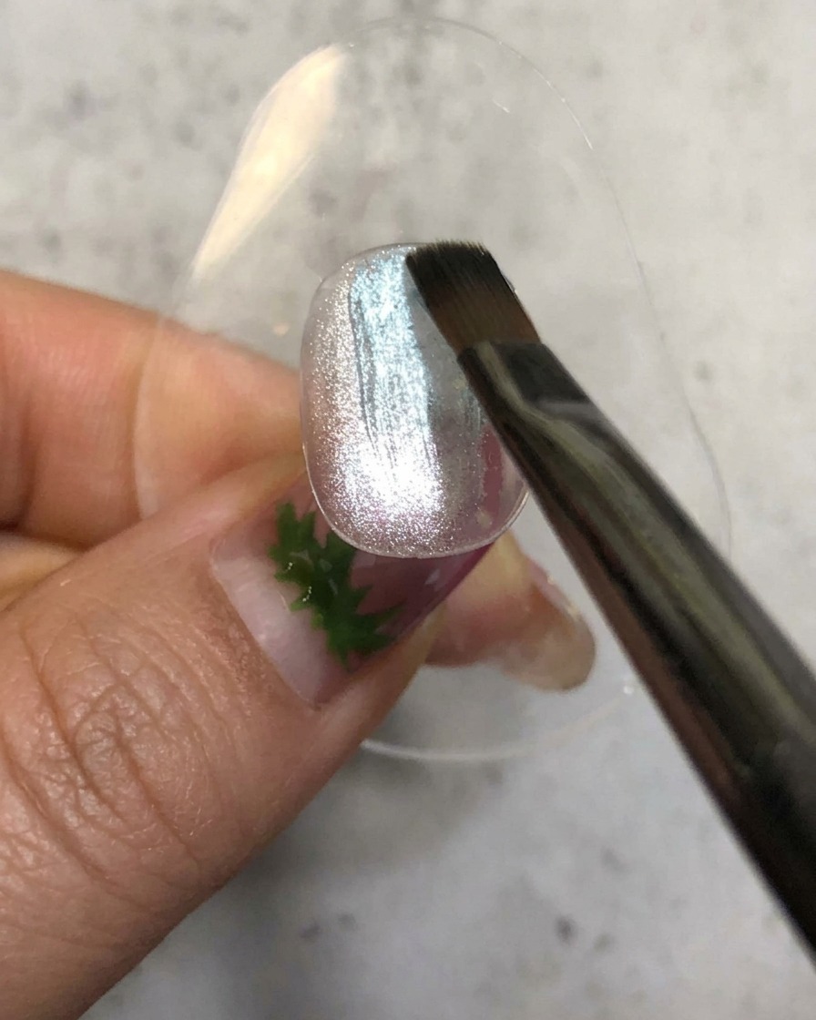 A nail brush applying a sheer blue gel polish over a sparkly silver magnetic base.