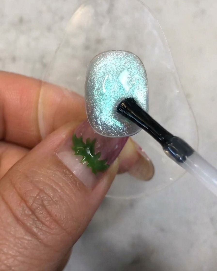 A brush applying a small drop of clear reinforcement gel onto the textured blue cat-eye nail surface.