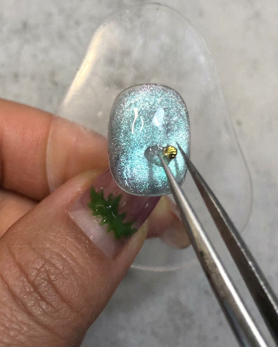 A pair of tweezers placing a tiny golden seashell charm onto the reinforcement gel dot on a blue nail.