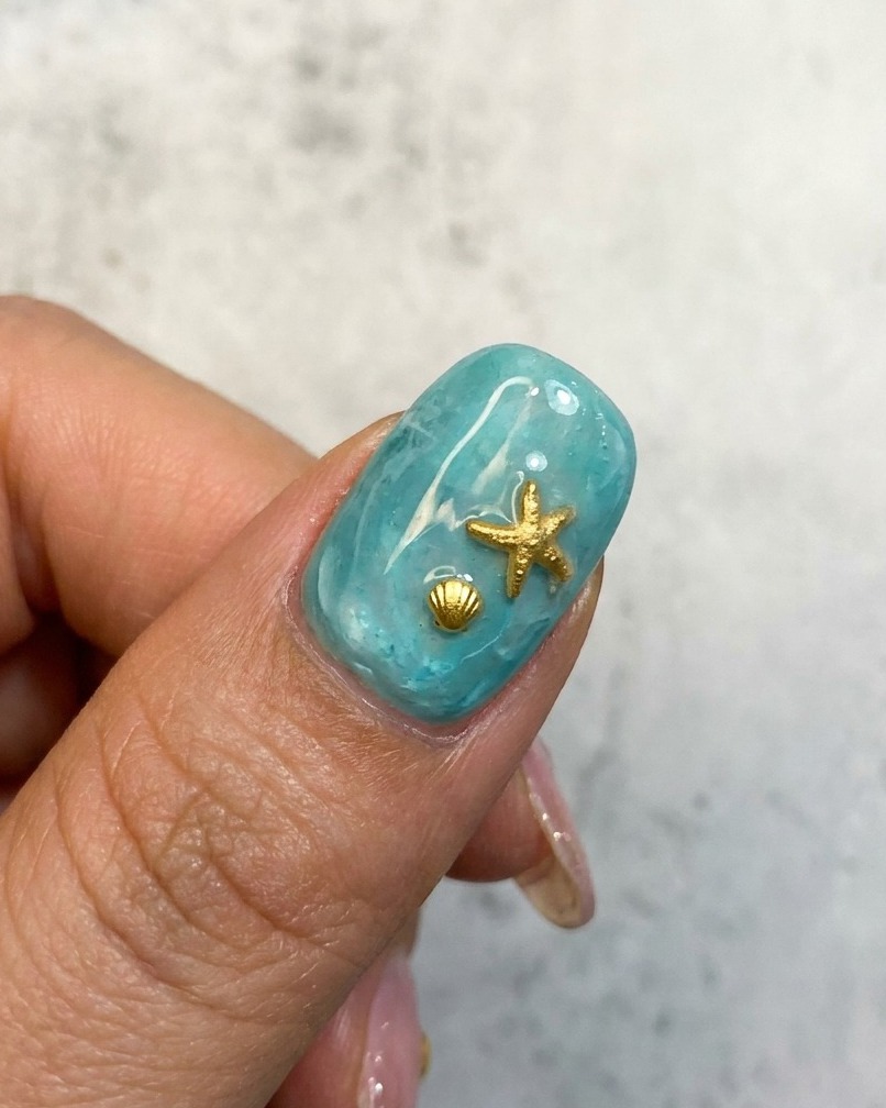 A close-up of a blue oceanic nail design with a gold starfish and seashell embellishment.