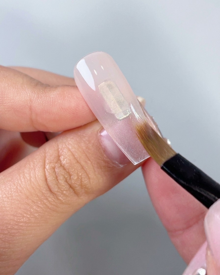 A close-up of a flat makeup brush blending translucent pink gel downwards on a clear artificial nail tip.