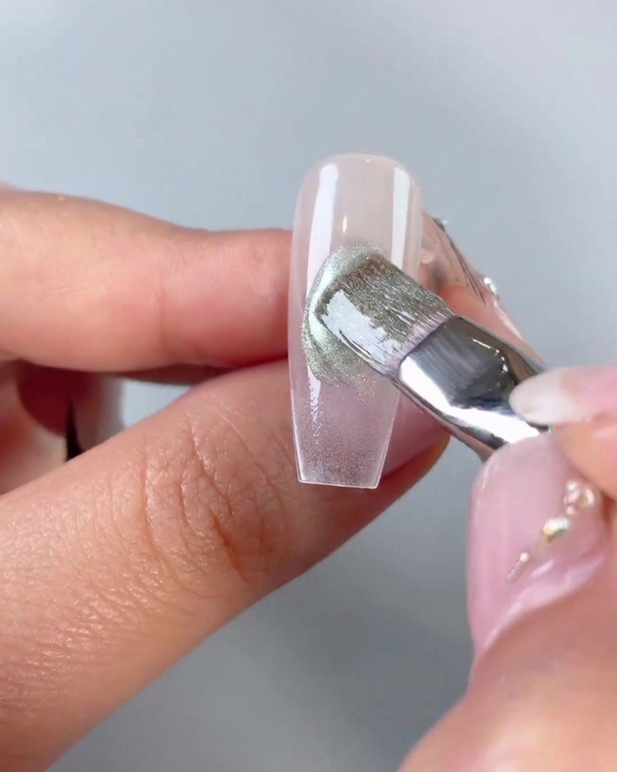 A flat brush smoothing and blending green cat-eye gel applied diagonally across the center of a clear nail tip.