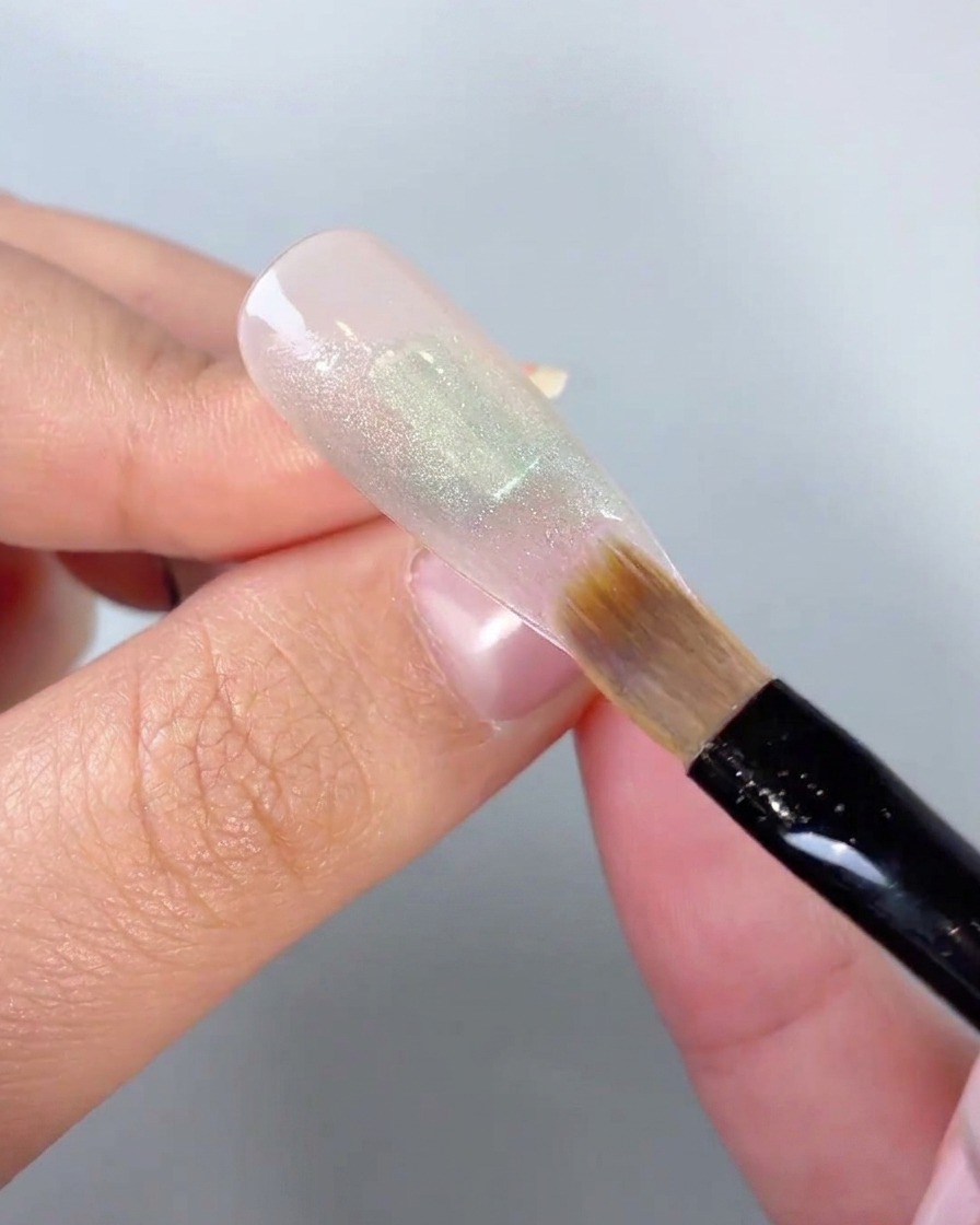 A close-up of a soft brush blending the edges of a green gel color block on a nail tip.