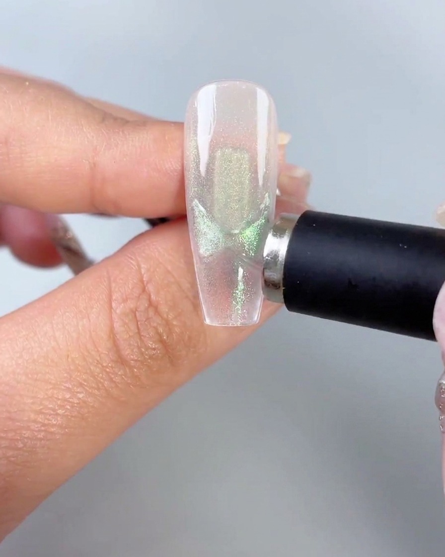 A black magnetic wand held close to the side of a clear nail tip showcasing activated green cat-eye butterfly wing patterns.