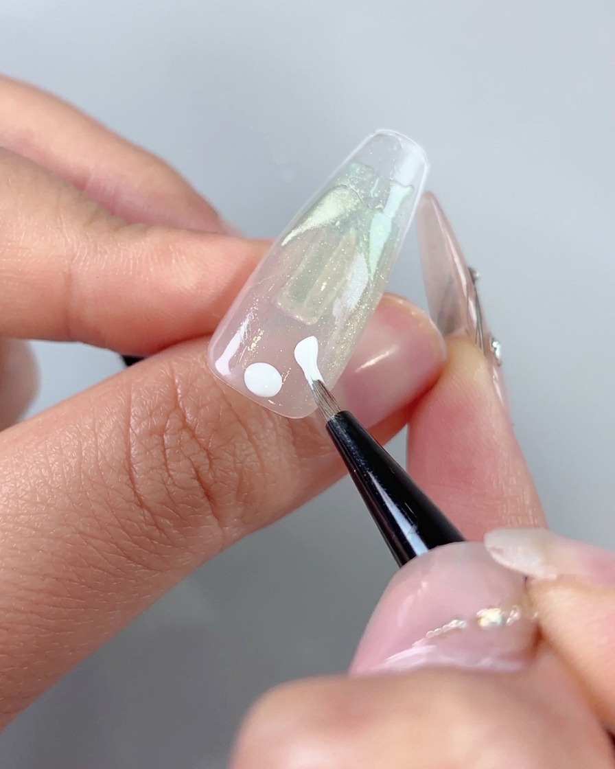A fine art brush applying white gel dots and initial strokes near the cuticle area of a nail.