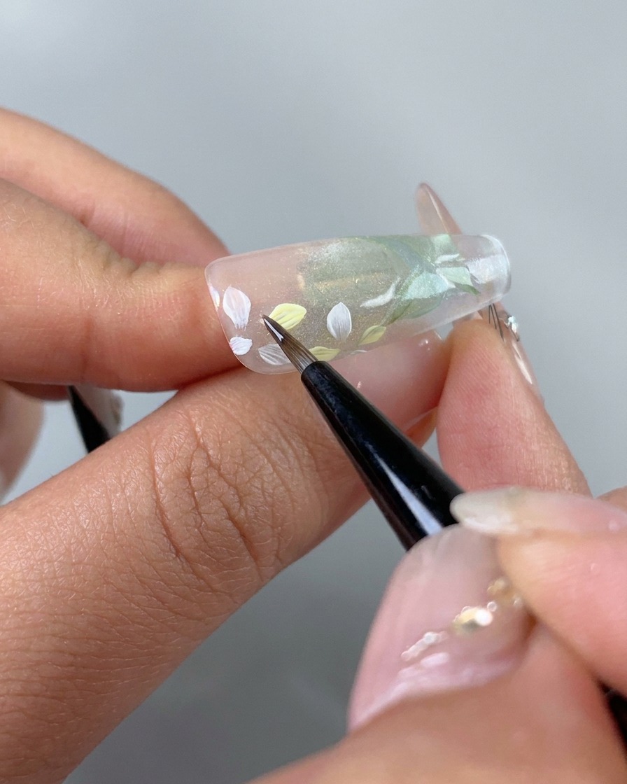 A fine brush carefully adding light pink and yellow gel tints onto individual white painted flower petals on the nail.