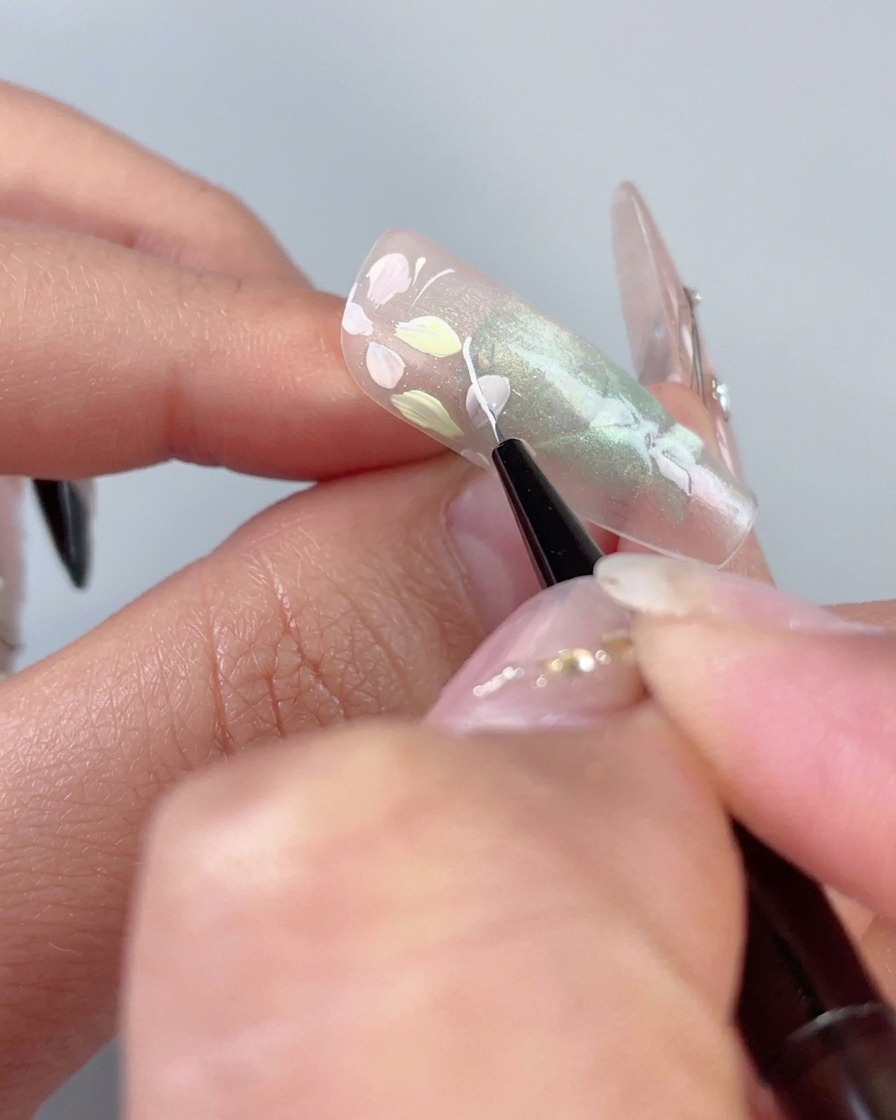 A fine liner brush drawing a delicate white curved line next to pastel flowers on a translucent green cat-eye nail.