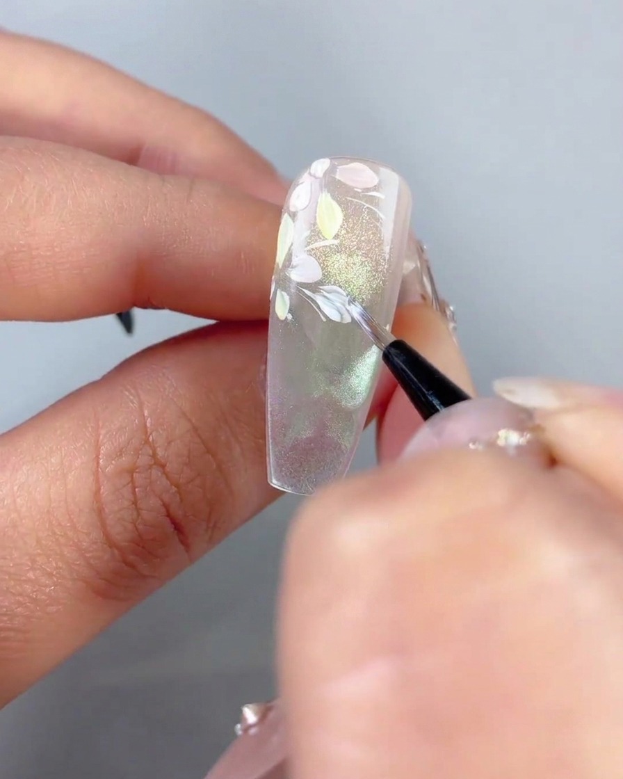 A fine brush dabbing tiny white leaf accents onto curved white lines near pastel flower petals on a nail.