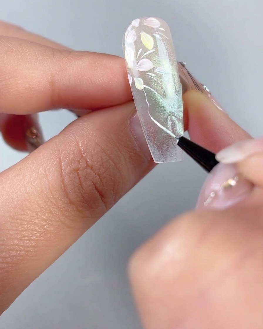 A fine liner brush tracing thin white outlines and wing veins over a sheer green cat-eye butterfly shape on a nail.