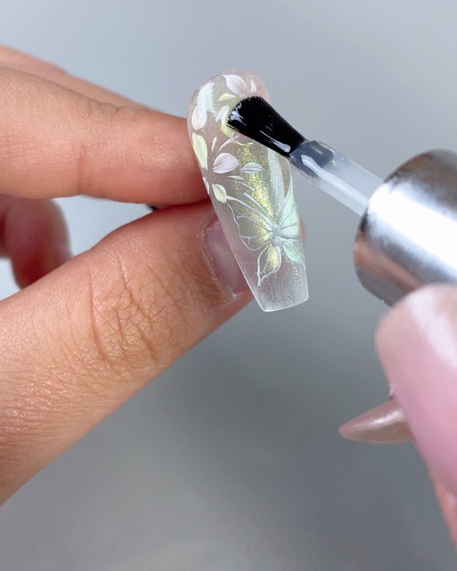 A nail polish brush applying a glossy clear top coat over a fully decorated translucent floral and butterfly nail.