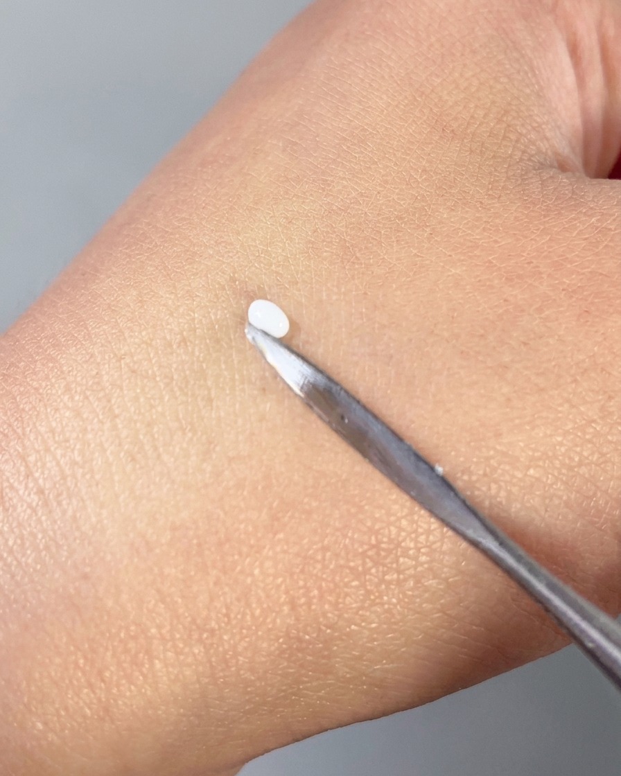 A metal spatula pressing a small white bead of 3D gel into a smooth petal shape on the back of a hand.