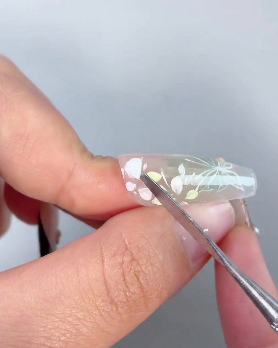 A metal tool applying a drop of clear adhesive gel onto the center of a painted floral design on a nail.