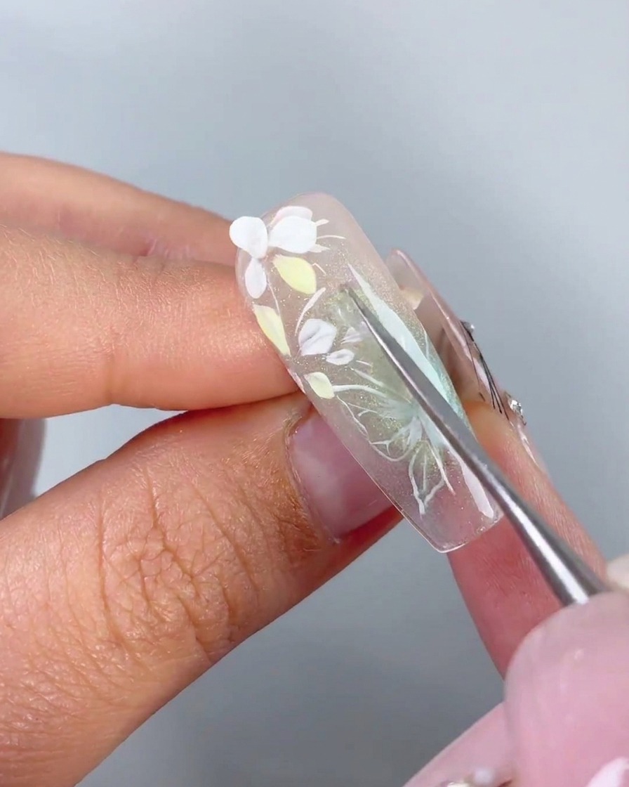 A metal tool placing a pre-shaped white 3D petal onto the adhesive gel of a nail.