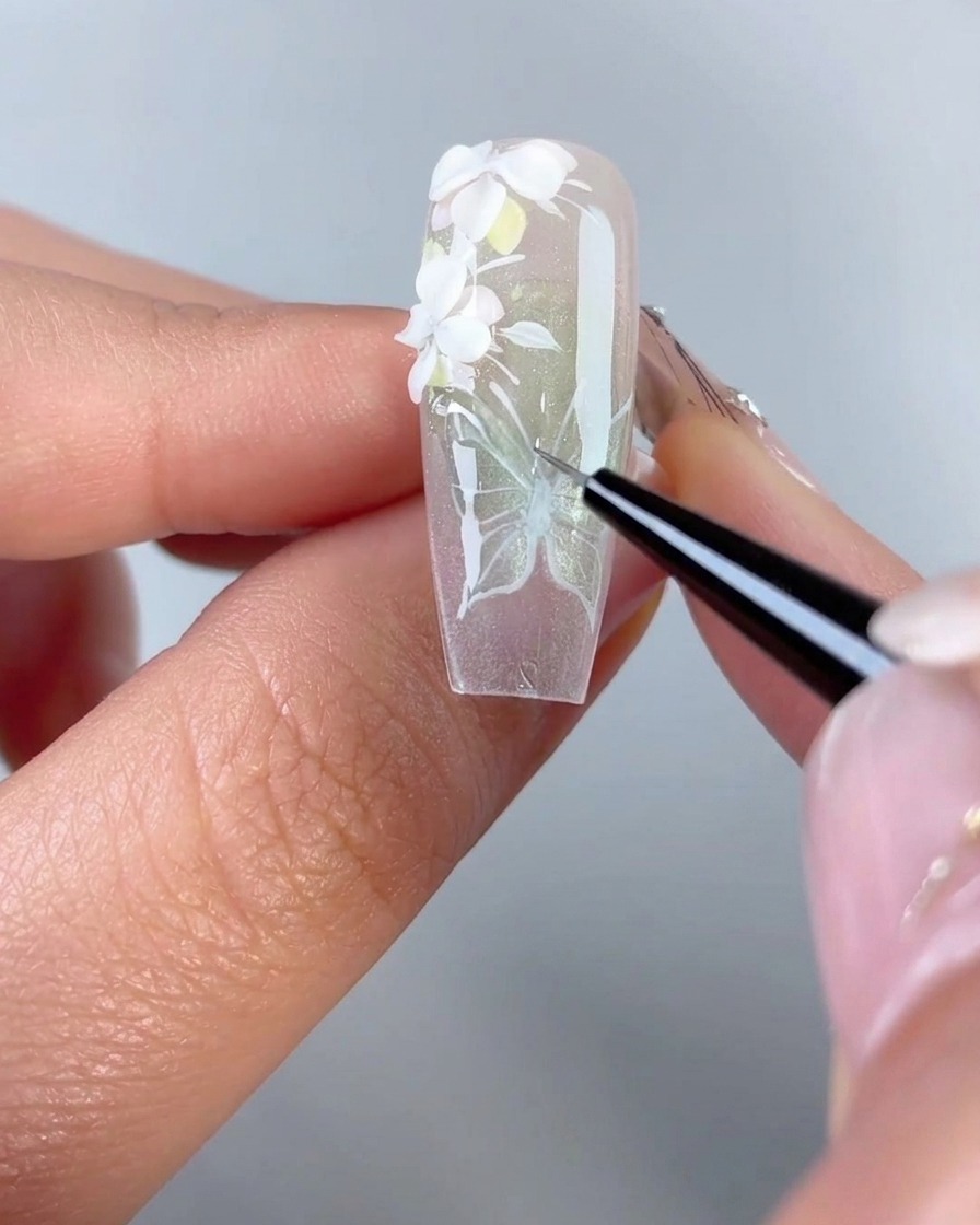 A close-up of a fine liner brush tracing clear 3D modeling gel over the white painted lines of a butterfly wing design on a gel nail tip.