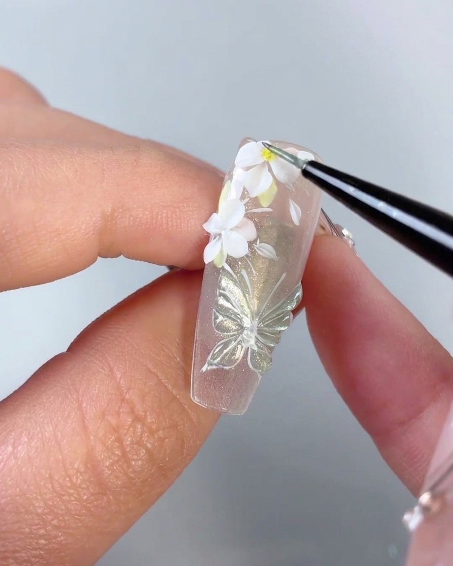 A fine brush adding a tiny yellow dot to the center of a white 3D flower on a nail.