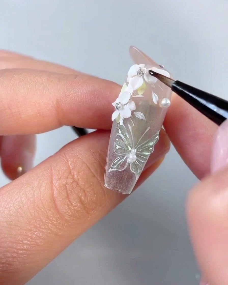 A fine detail nail brush applying clear gel around the edges of white 3D flower petals and rhinestones on a clear nail tip.