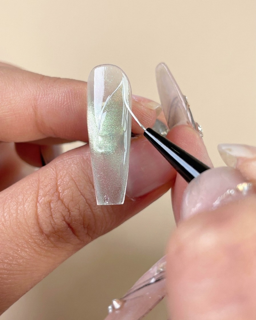 A fine liner brush drawing white vein lines on a green cat-eye gel nail.