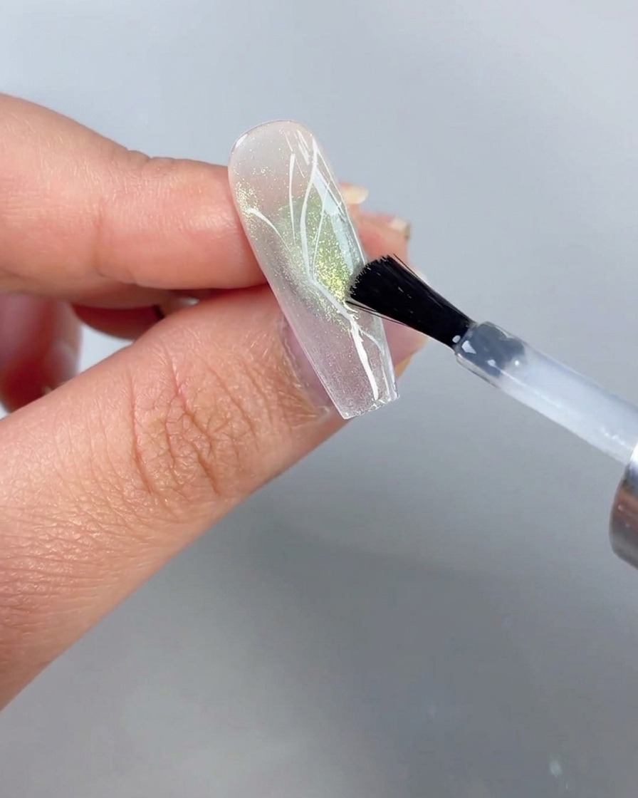 A brush applying clear top coat over a nail featuring a painted half-butterfly design against a sheer green cat-eye background.