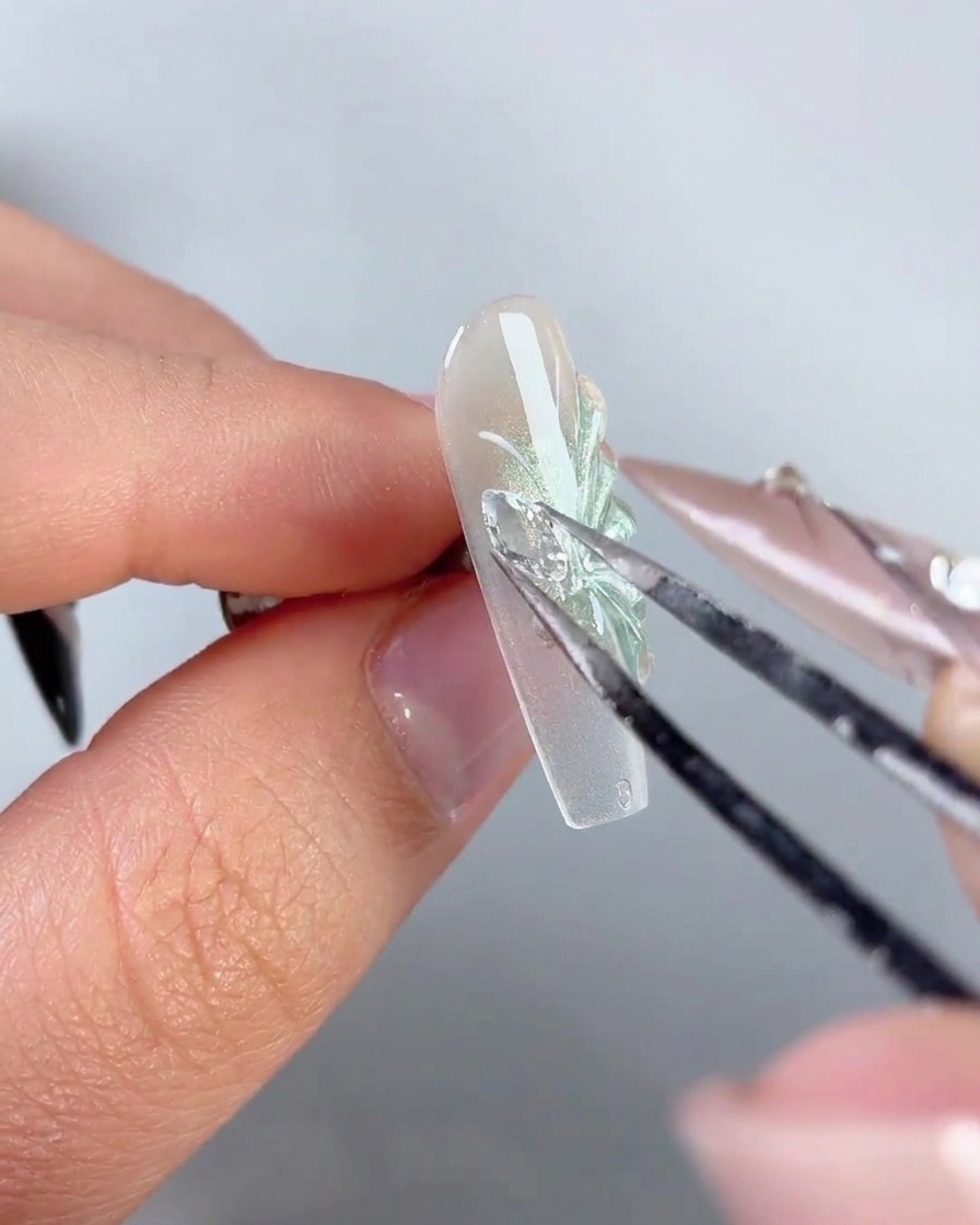 A pair of metal tweezers placing a sparkling rhinestone butterfly wing charm onto the side of a decorated nail.