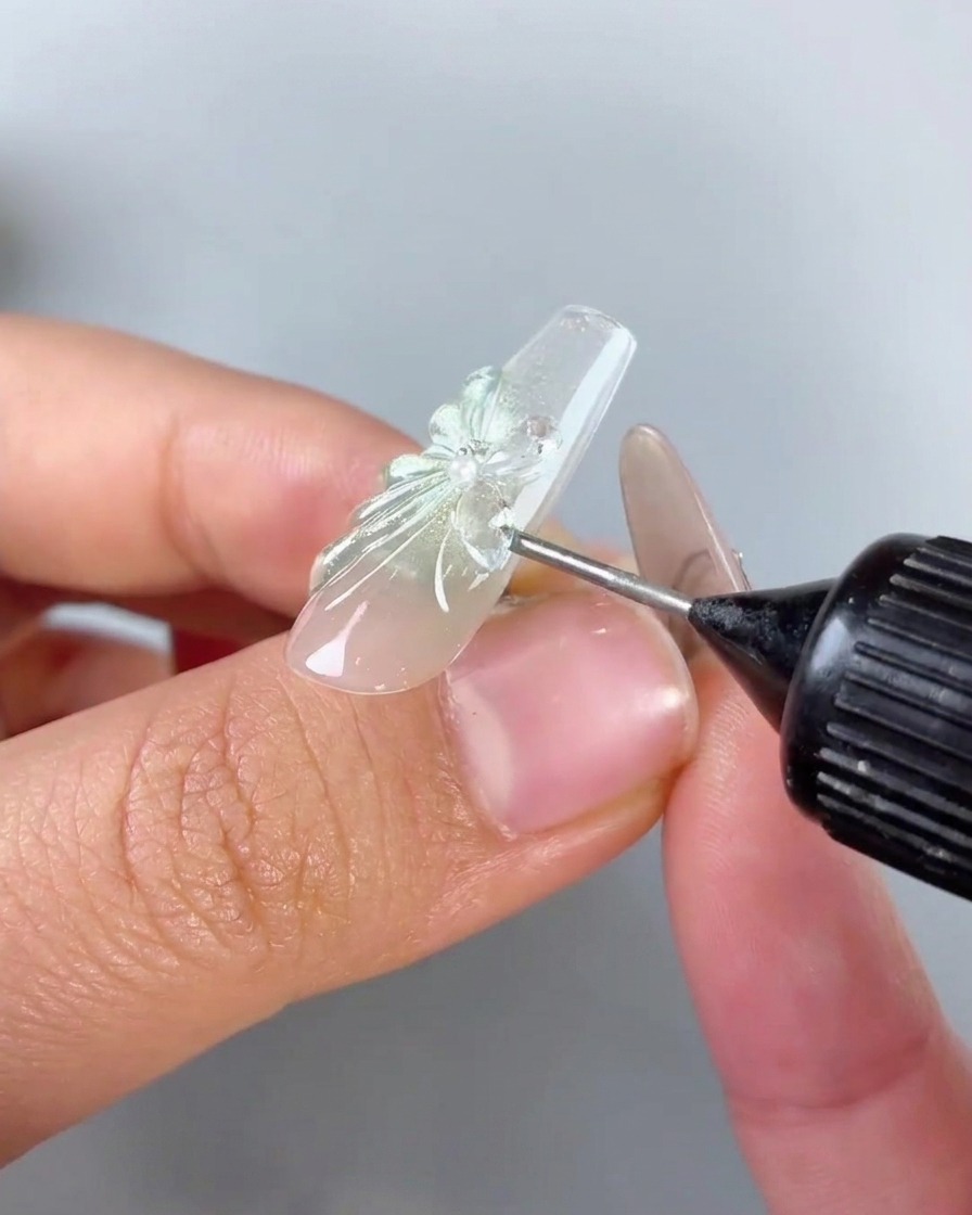 A needle-tip squeeze bottle applying clear gap-filling gel around the base and edges of a rhinestone butterfly charm on a nail.