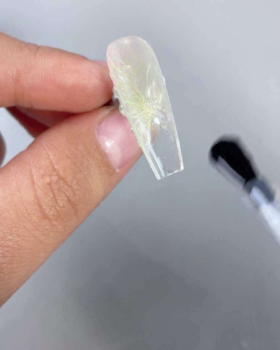 A close-up of a hand holding an ice-transparent nail with a 3D floral design upside down, while a brush applies clear top coat to the underside of the free edge.