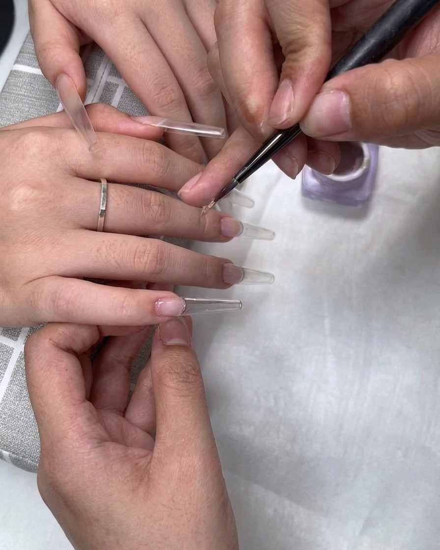 A fine brush applying milk tea nude gel polish near the cuticle of a clear nail extension.