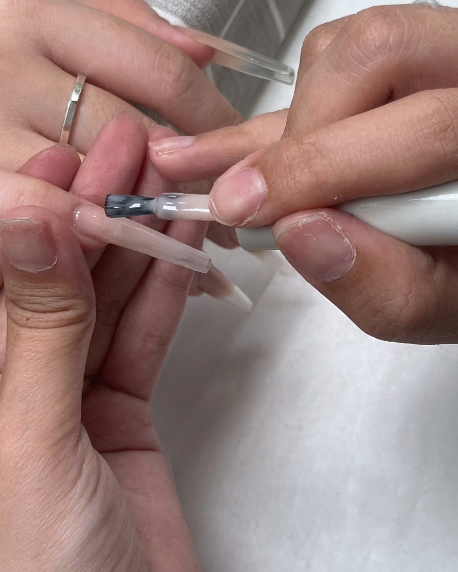 A brush applying a layer of sheer nude polish over the entire nail surface.