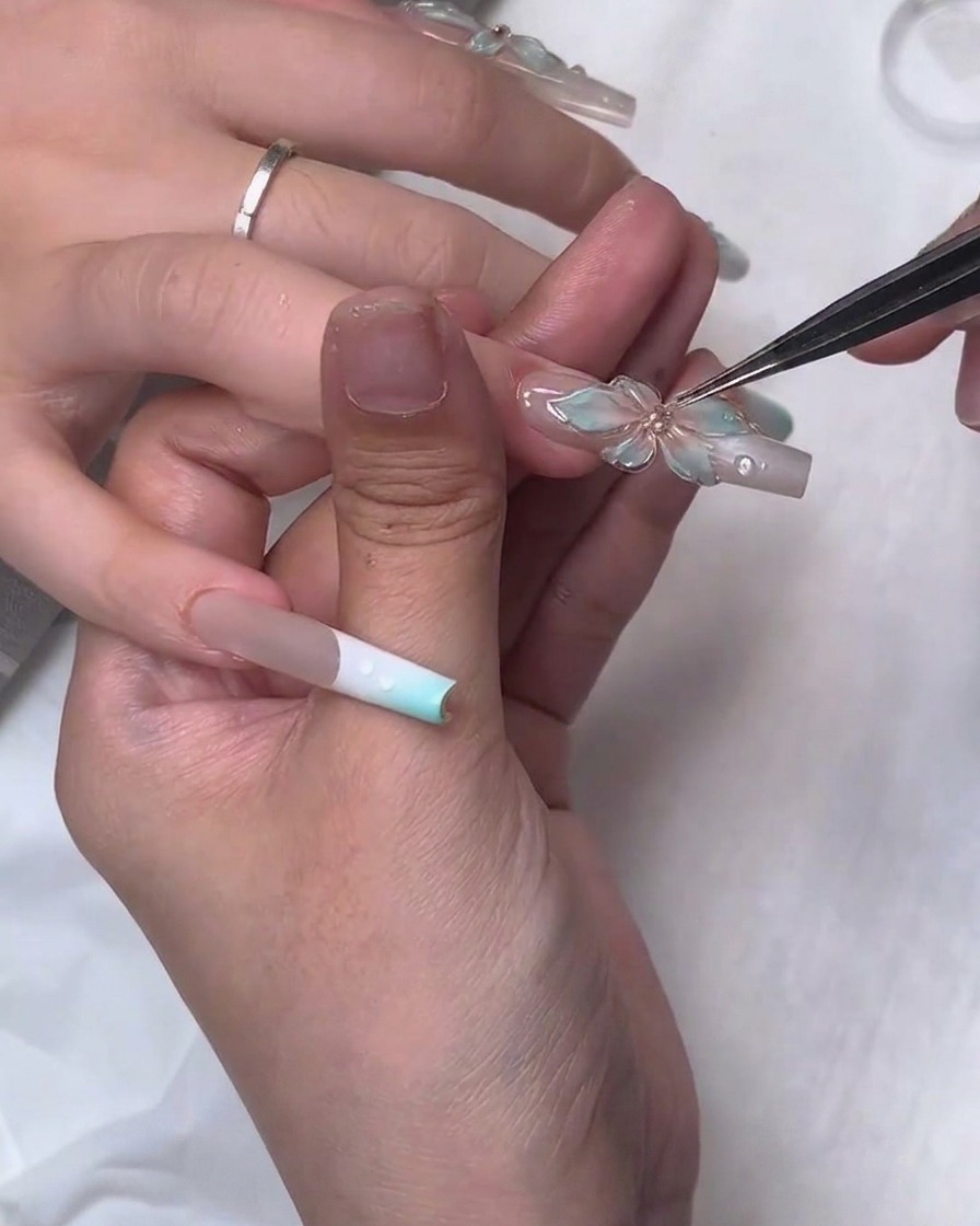 A close-up showing fine tweezers precisely placing a small rhinestone jewel into the center of a translucent 3D sculpted gel flower on a long square fingernail.