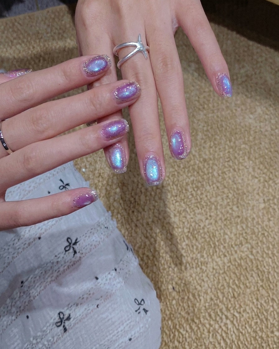 A brush applying a coat of shimmering blue-purple cat eye gel polish onto a nail.