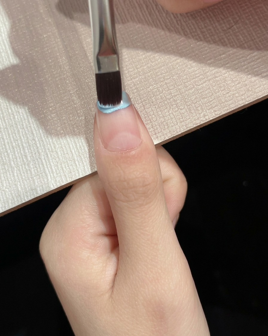 A flat brush applying light blue gel polish onto the upper half of a nail plate.