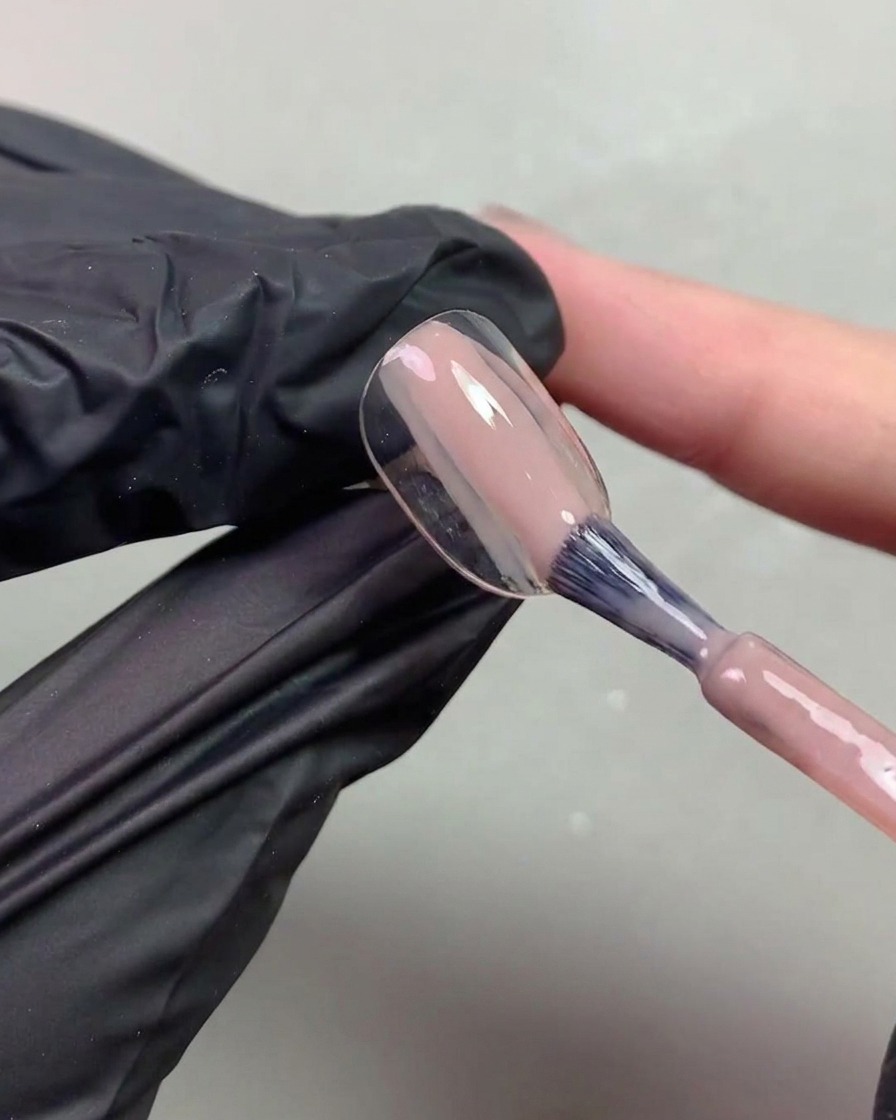 A close-up of a hand in a black glove using a brush to apply a sheer nude orange-toned gel to a nail tip.