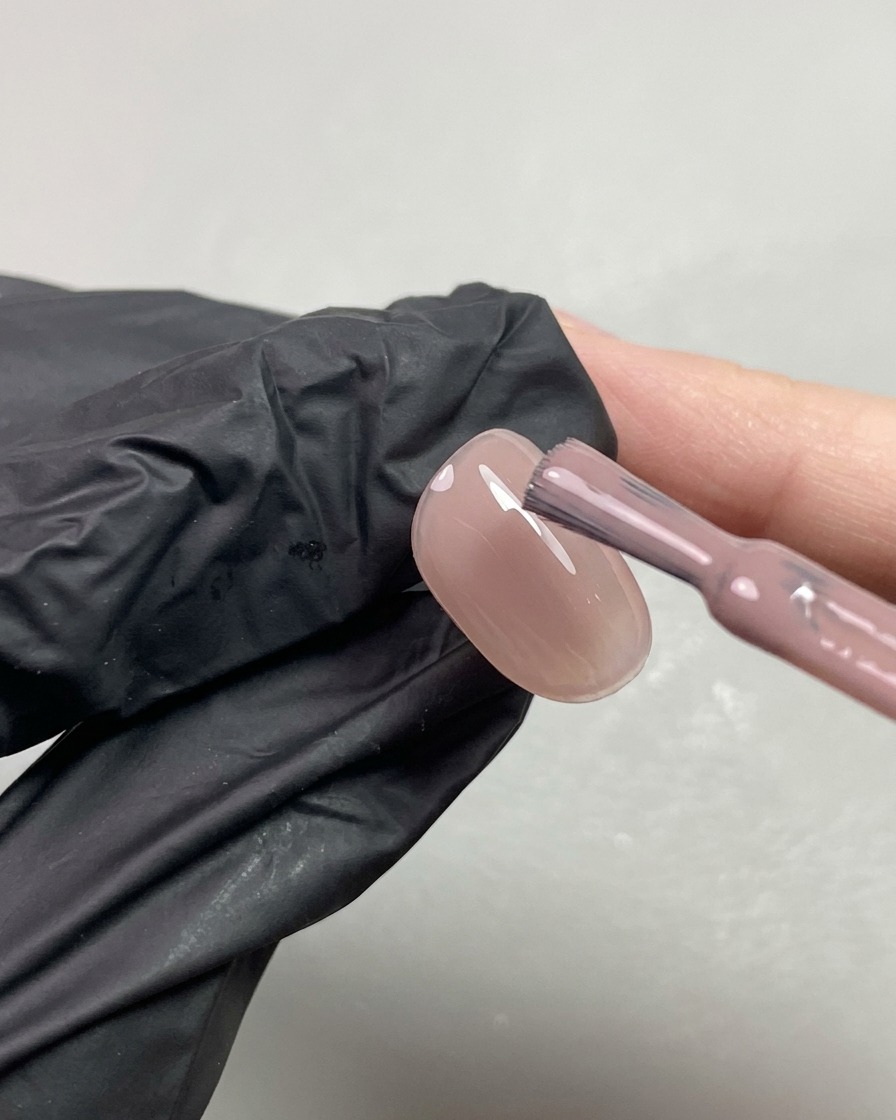 A brush applying a second coat of sheer nude orange-toned gel to a fingernail.