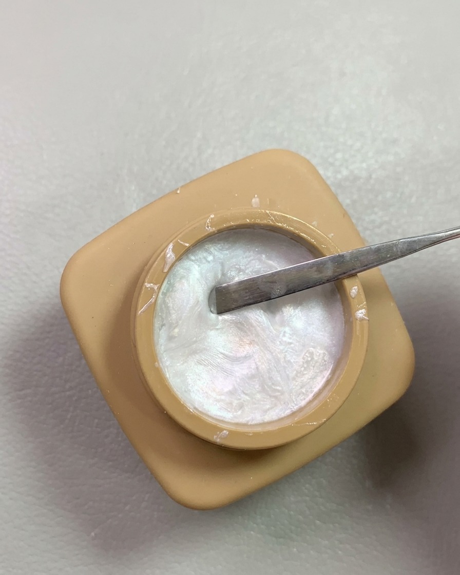 A metal spatula tool scooping shimmering nacreous pearl carving gel from a small tan jar.