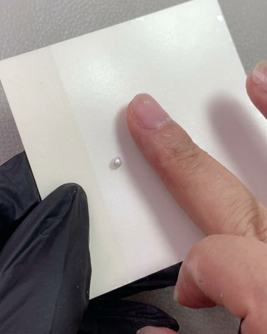 A gloved hand pointing to a small nacreous pearl carving gel ball placed on a piece of mixing paper.