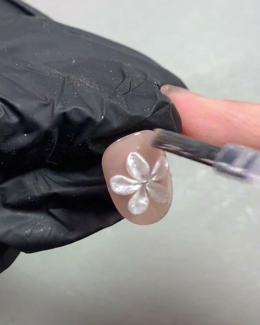 A gloved hand placing a short nail adorned with a 3D white pearl flower under a purple UV/LED curing lamp.