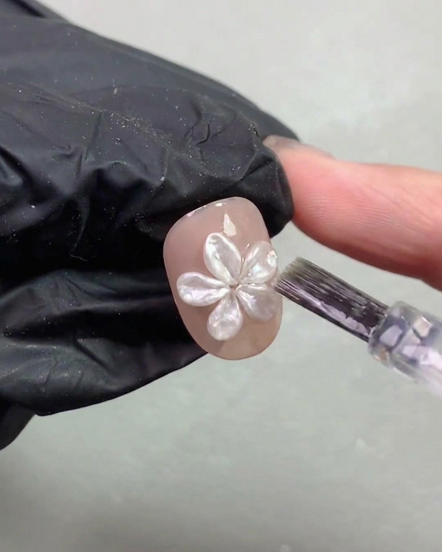 A nail brush carefully applying a thin top coat layer around a cured 3D pearl flower on a nail.
