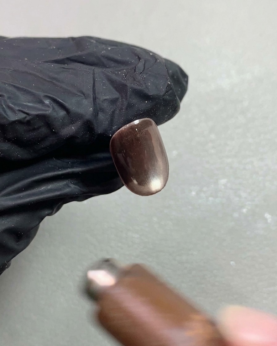 A nail magnet hovering above a short dark brown cat-eye gel nail, shaping the reflective pigment into a bright rolling sphere effect.