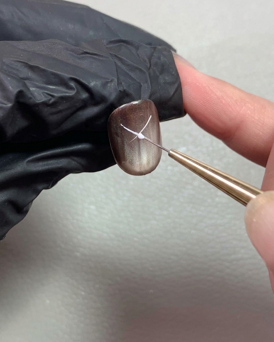 A fine liner brush drawing thin white lines to start forming a bowknot on a dark brown glossy nail.