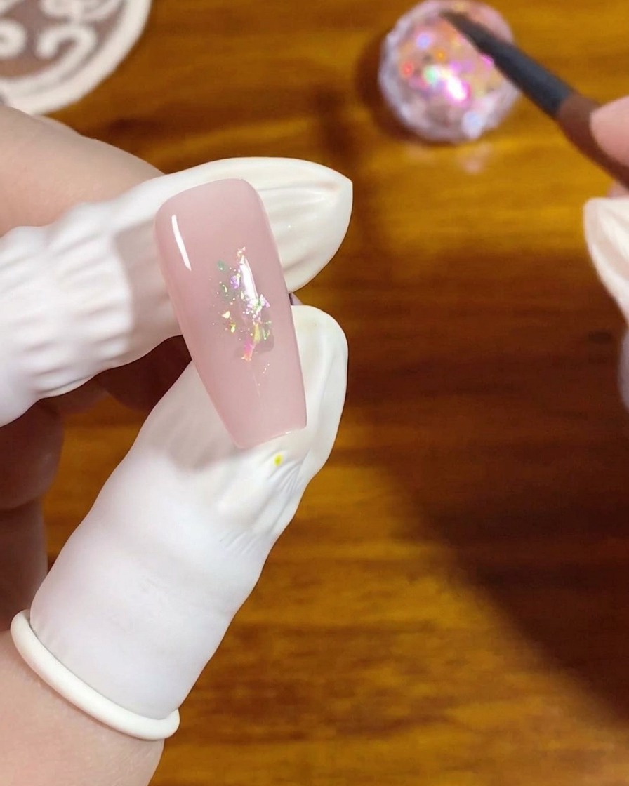 A flat gel brush pressing small iridescent opal flakes onto the center of a pink polished nail.