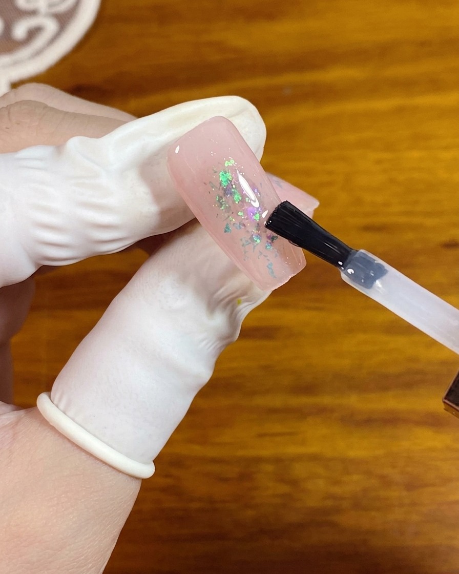 A gel brush applying a clear matte top coat over a pink nail with opal flakes in the center.