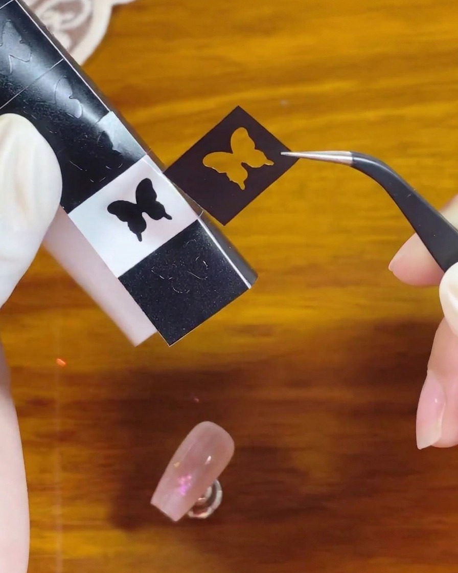 Tweezers peeling a black butterfly silhouette stencil sticker from a sheet to place onto the nail.