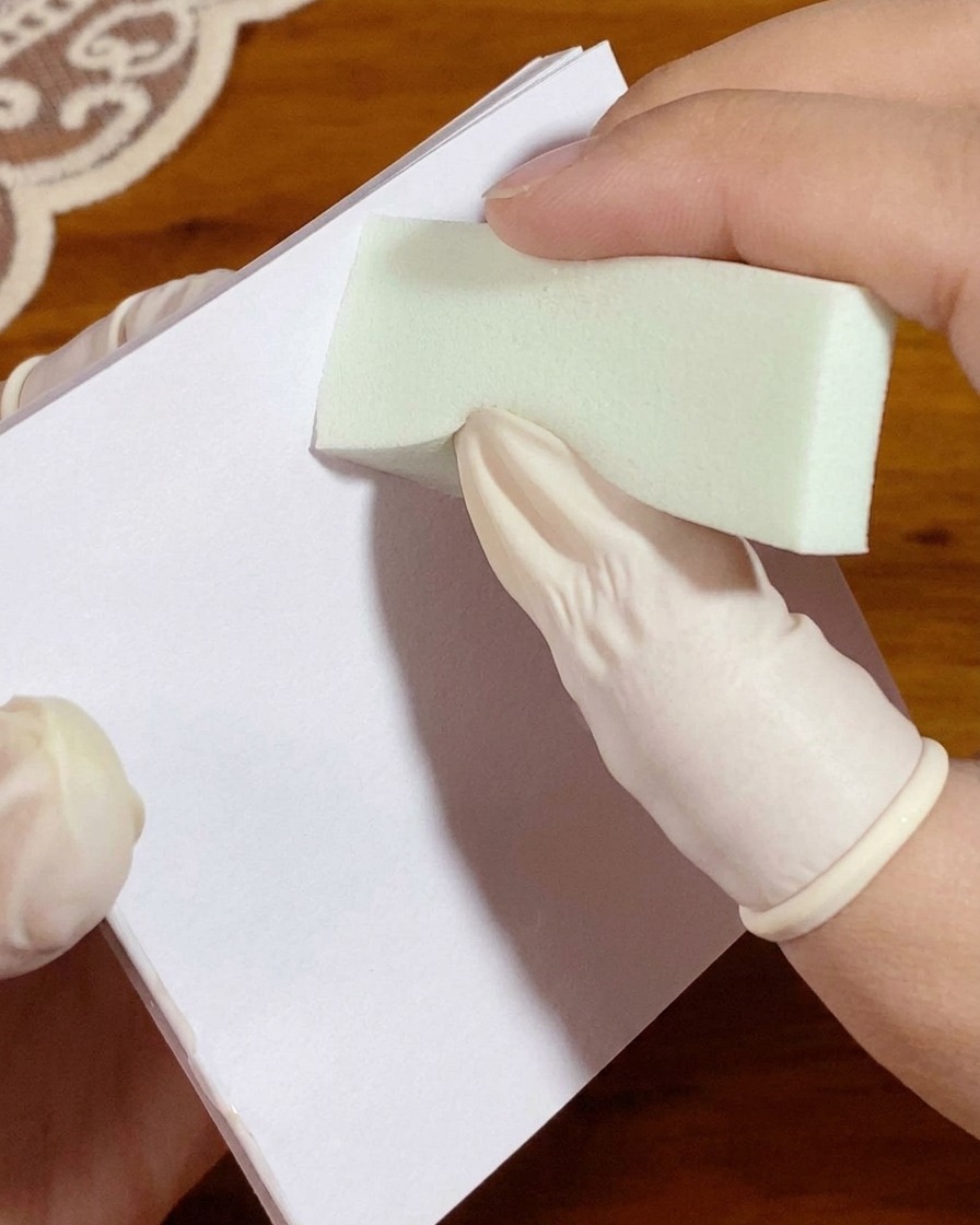 A gloved hand pressing a sponge loaded with white gel onto a palette paper before nail application.
