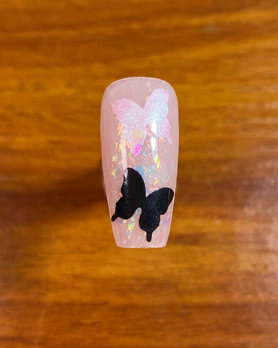A top-down view of a decorated nail featuring iridescent flakes and two butterfly stencil shapes.