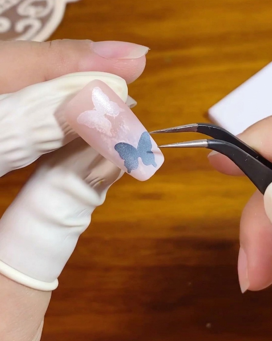 A pair of tweezers lifting a stencil sticker off a pink gradient nail design.